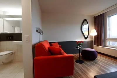 a living room with a red chair and a bathroom at Hotel Thurgauerhof Self-Check-in in Weinfelden